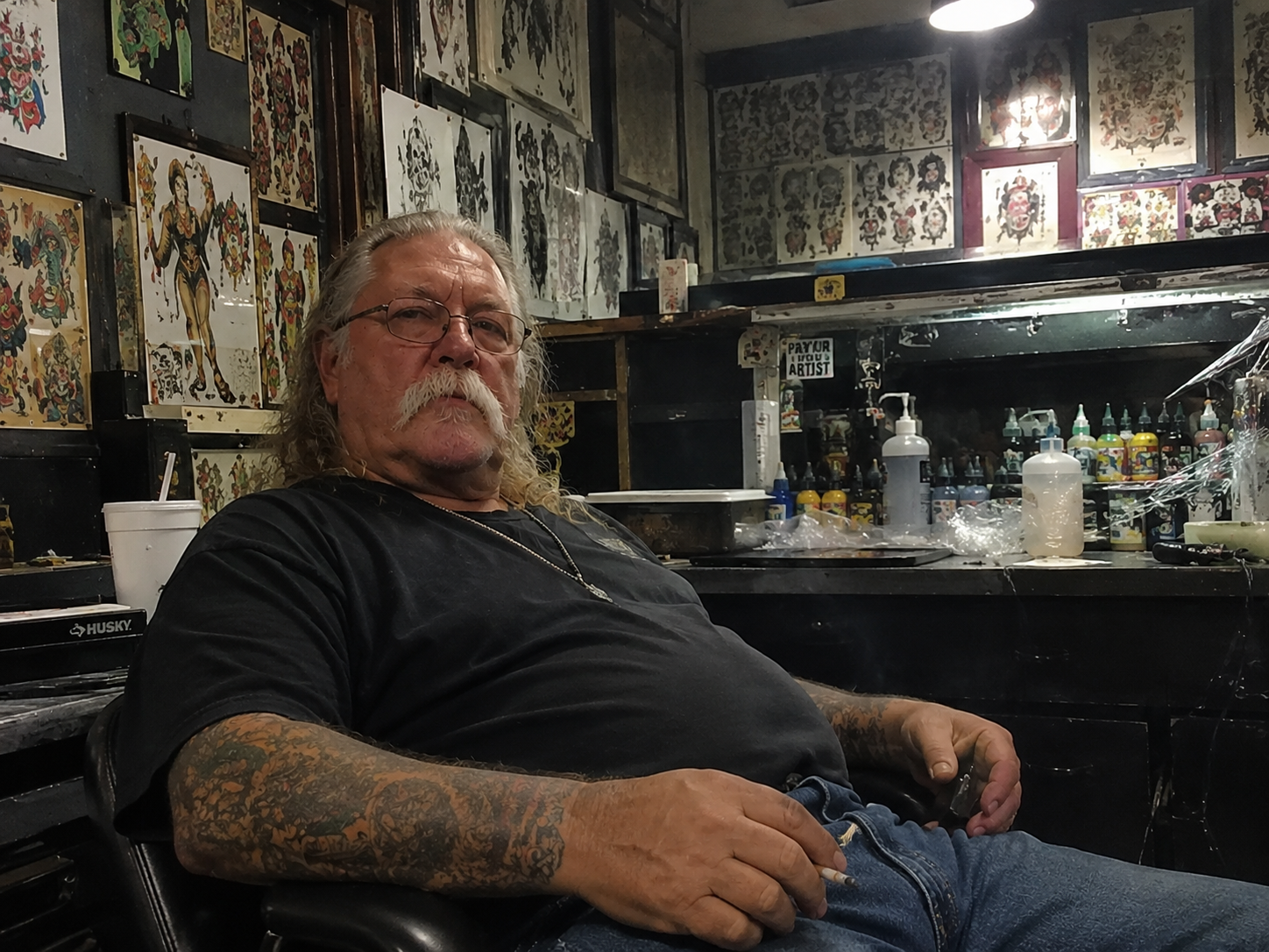 Old school tattoo artist seated inside a tattoo shop surrounded by flash sheets and tattoo equipment